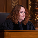 Image of a female judge with long, red, curly hair, wearing a black judicial robe seated at a wooden courtroom bench.