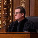Image of a male judge wearing a black judicial robe sitting at a wooden bench in the courtroom of the Thomas J. Moyer Ohio Judicial Center.