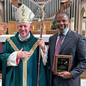A white Catholic bishop standing next to a Black man in a suit and tie with an award.