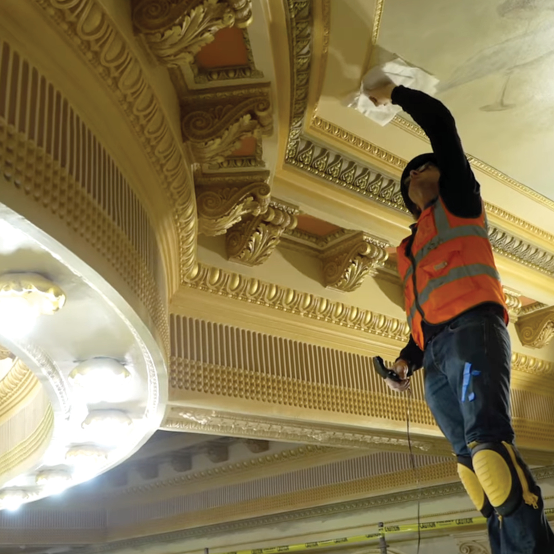A man wearing an orange safety vest working on an ornate ceiling.