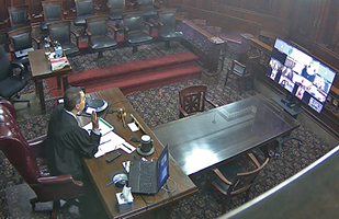 Image of a judge sitting at a desk in an empty room conducting adoption proceedings via video conferencing