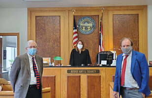 Image of two men standing on either side of a courtroom bench and a female judge standing behind the bench - all wearing protective masks
