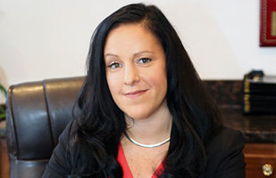 Image is of judge-designate  Courtney Caparella-Kraemer sitting in a leather chair at a desk, wearing a dark suit and orange blouse