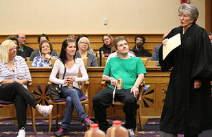 Image of about a dozen people sitting in a courtroom gallery listening to a woman judge who is in her black judicial robe