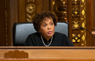 Black woman with curly hair wearing a pearl necklace and judidical robe sitting behind a bench.