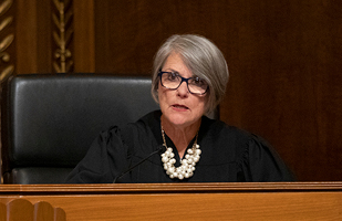 Woman with glasses and grey hair in a black judicial robe sitting behind a bench.