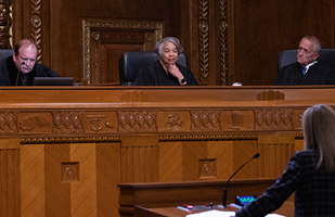 Image of a female judge wearing a black judicial robe seated between two males judges, also wearing black judicial robes. A woman stands in front of them at a podium.