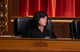 Image of a female judge with brunette hair wearing a black judicial robe seated at a wooden bench in the courtroom of the Thomas J. Moyer Ohio Judicial Center.