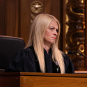 Image of a female judge with blonde hair wearing a black judicial robe seated at a wooden bench in the courtroom of the Thomas J. Moyer Ohio Judicial Center.