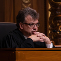 Image of a male judge with short, brunette hair wearing glasses and a black judicial robe sitting at a wooden bench in the courtroom of the Thomas J. Moyer Ohio Judicial Center.