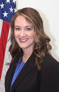 A woman with long, brown hair wearing a black suit jacket and blue blouse standing in front of an American flag.