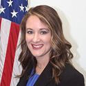 A woman with long, brown hair wearing a black suit jacket and blue blouse standing in front of an American flag.