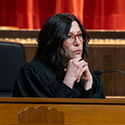 A woman with long, dark hair, wearing glasses and a black judicial robe seated behind a wooden bench in the courtroom of the Thomas J. Moyer Ohio Judicial Center.