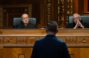 A man wearing a black judicial robe is seated at a wooden bench looking at a man wearing a blue suit standing in front of the bench in the courtroom of the Thomas J. Moyer Ohio Judicial Center.