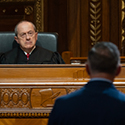 A man wearing a black judicial robe is seated at a wooden bench looking at a man wearing a blue suit standing in front of the bench in the courtroom of the Thomas J. Moyer Ohio Judicial Center.