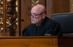 A male judge wearing a black judicial robe and glasses seated at a wooden bench in the courtroom of the Thomas J. Moyer Ohio Judicial Center