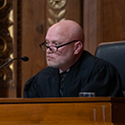 A male judge wearing a black judicial robe and glasses seated at a wooden bench in the courtroom of the Thomas J. Moyer Ohio Judicial Center.