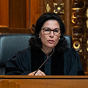 A woman with shoulder-length, dark hair and glasses, wearing a black judicial robe sits at a wooden bench in the courtroom of the Thomas J. Moyer Ohio Judicial Center.