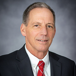 A man wearing a navy blue blazer, white dress shirt, and red tie.