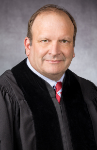 A man wearing a black judicial robe, white dress shirt, and red striped tie.