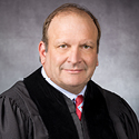 A man wearing a black judicial robe, white dress shirt, and red striped tie.