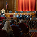 A woman speaking at a podium with a portrait to an audience of men and women