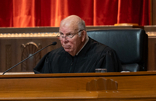 A man sitting at a bench speaking into a microphone wearing judicial robes.