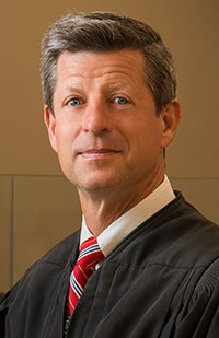 A man wearing a black judicial robe, white dress shirt, and red striped tie.