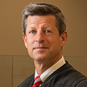 A man wearing a black judicial robe, white dress shirt, and red striped tie.