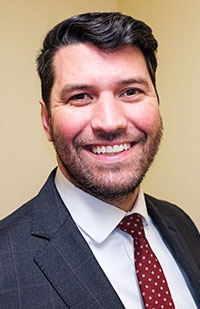 A man with short, dark hair and a beard wearing a blue checked blazer, white shirt, and red tie.