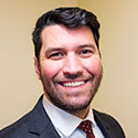 A man with short, dark hair and a beard wearing a blue checked blazer, white shirt, and red tie.