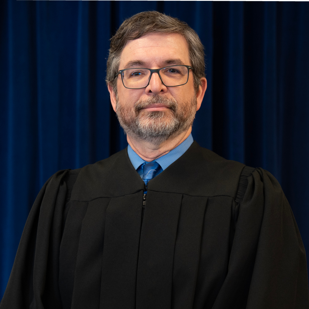 Three judges wearing black robes stand against a blue curtain backdrop. Each person is centered in their own segment of the arrangement.
