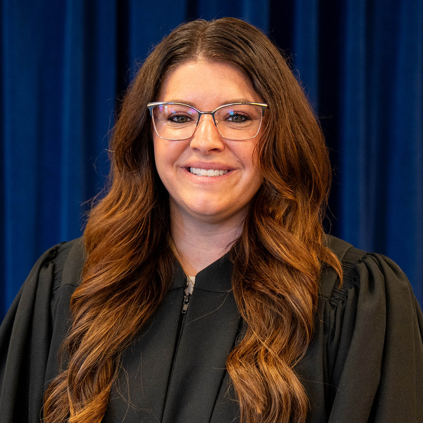 A woman with long, wavy brown hair wearing glasses and a black robe smiles in front of a blue curtain.