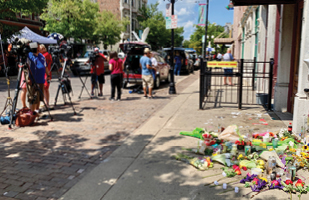 Image of a sidewalk with a memorial of flowers outside a building and television camera crews set up in the street (flickr/Scott Beale)