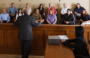 Image of an attorney addressing 12 jury members, with a man sitting at a table next to him