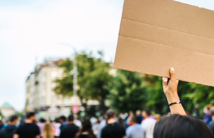 Image of a woman in a crowd of people holding up a cardboard sign in protest (iStock/<a href='https://www.istockphoto.com/portfolio/VasilDimitrov?mediatype=photography'>Vasil Dimitrov</a>)
