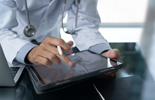 Image of a male doctor wearing a long, white coat and a stethoscope around his neck. He is holding a stylus and has a tablet in his hand.