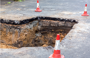 Image of a large sinkhole in pavement with orange construction cones on either side.