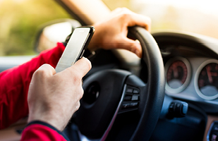 Image showing a person's hand holding a cell phone while the other hand is on a steering wheel.