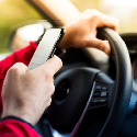 Image showing a person's hand holding a cell phone while the other hand is on a steering wheel.