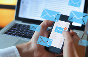 Image showing human hands holding a smart phone that has a large blue envelope on its screen. There are several large blue envelopes floating around the phone. There is a laptop in the background.