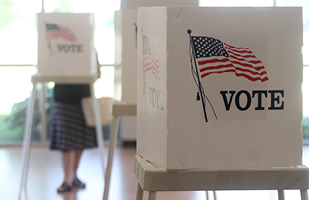 Image of a white privacy screen on top of a small table. The screen has an American flag and the word 'Vote' on it. In the background, a woman wearing a skirt stands behind a privacy screen voting.