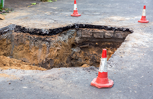 A very large hole in pavement. There are two orange construction cones on either side of the hole.