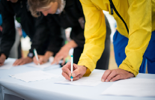 People leaning over a table and writing on a document with a pen.