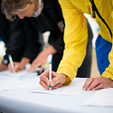 People leaning over a table and writing on a document with a pen.