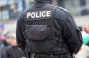 A police officer wearing black tactical uniform and gear stands with his back to the camera.