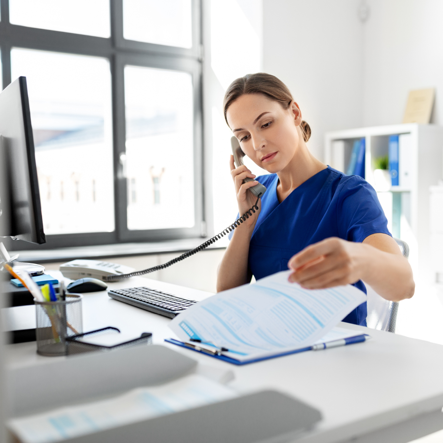 An administrative healthcare worker in blue scrubs sits at a bright office desk, speaking on a landline phone while reviewing a document on a clipboard.