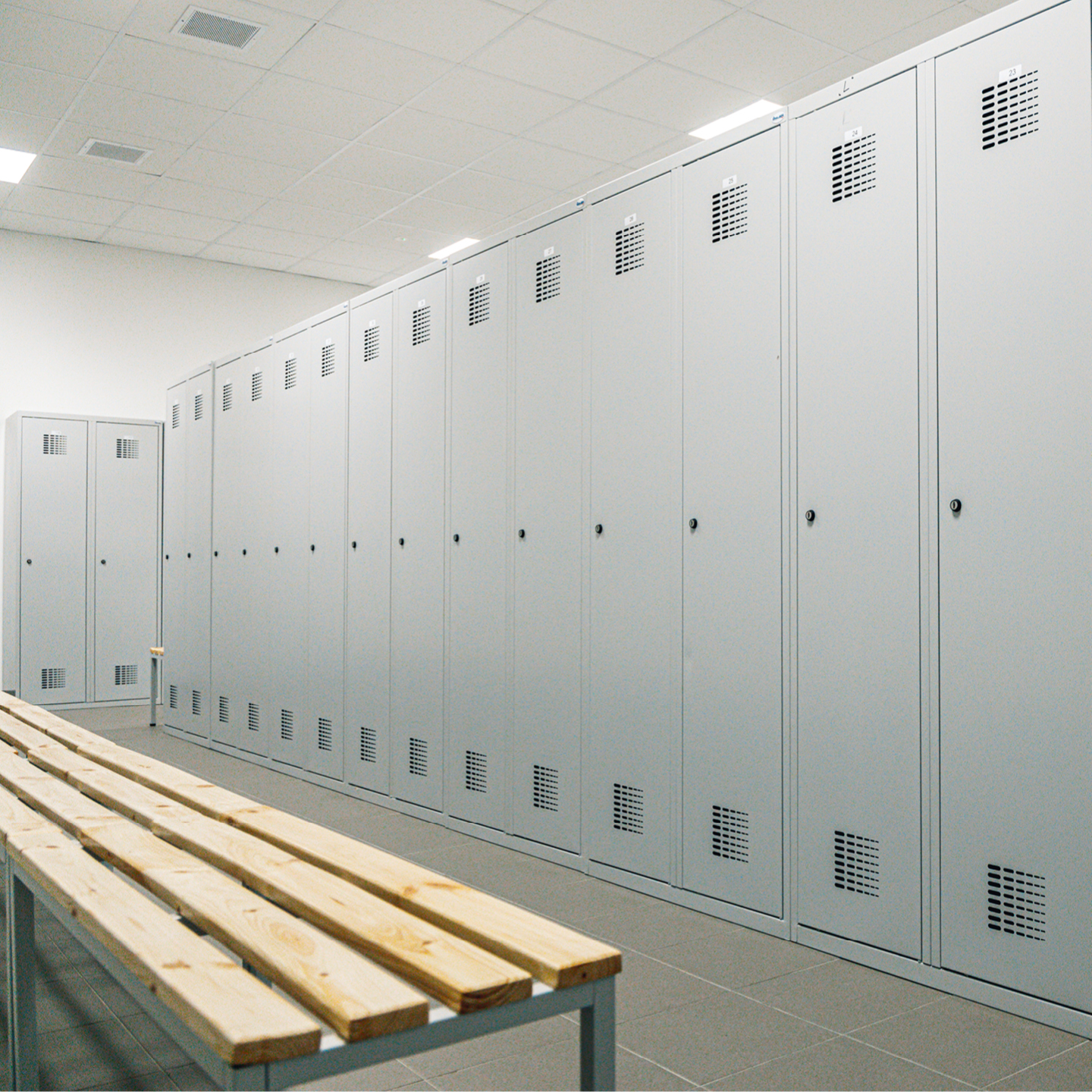 A long row of tall, light gray metal lockers lining a locker room wall next to a wooden bench.