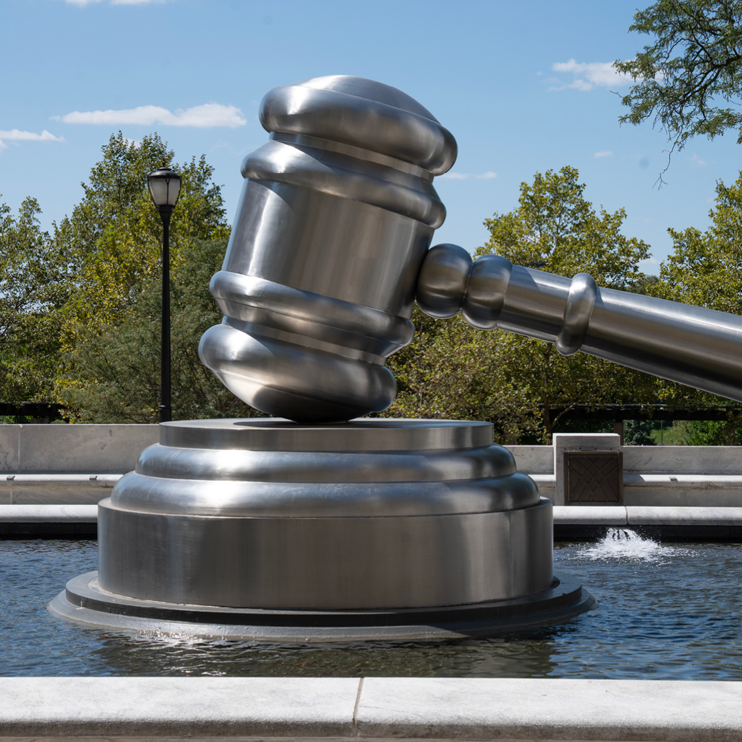 A large metallic gavel sculpture rests on its base in a fountain surrounded by trees and a blue sky.