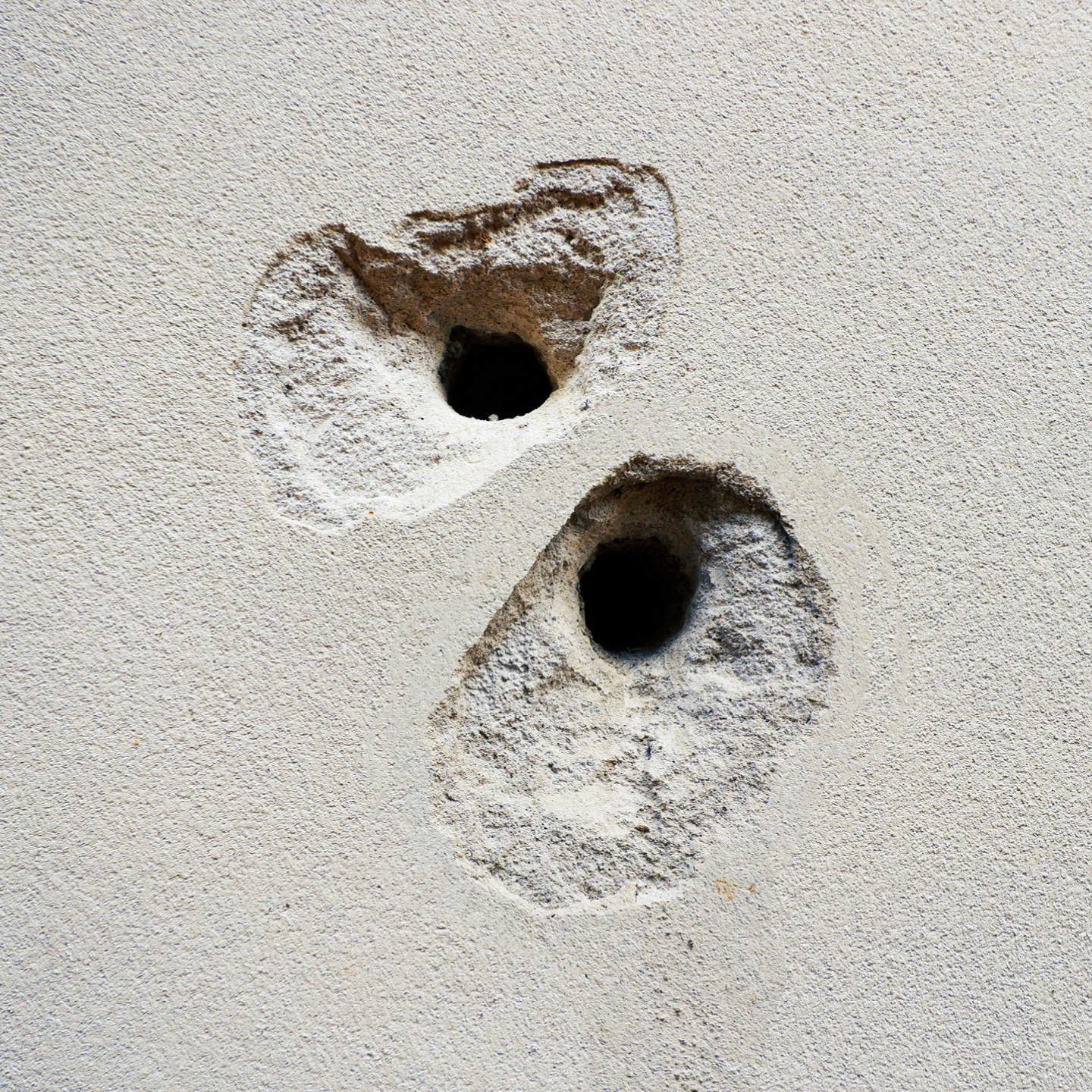 Two deeply gouged holes in a textured, light-colored concrete or stucco wall. The impact points feature dark, circular entry cavities in the center, surrounded by large, irregular areas where the outer surface material has chipped and crumbled away.